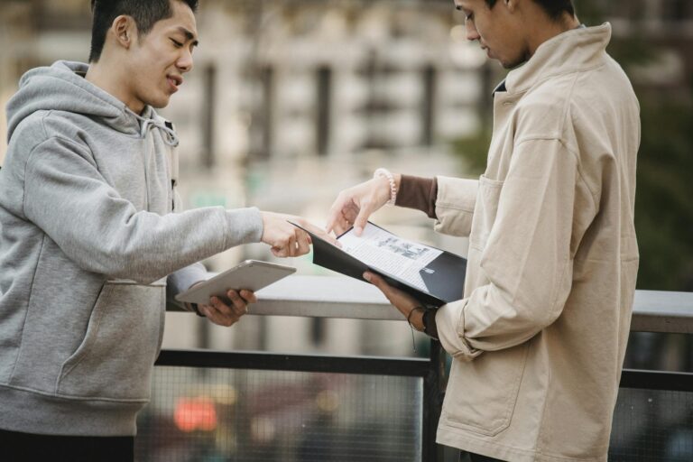 Side view of young diverse male students in casual clothes standing on street with tablet and papers in folder and speaking about home task